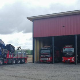 Neljä punaista Volvo-kuorma-autoa seisoo hallin edustalla tai sisällä. Yksi autoista on varustettu suurella sinisellä nosturilla ja pitkällä perävaunulla, kun taas kolme muuta ovat pysäköityinä hallin sisään. Halli on punainen ja harjakattoinen, ja ympärillä on sora-alusta sekä vihreää maisemaa taustalla.