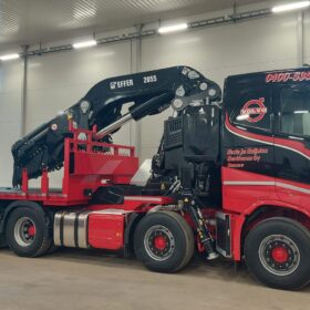 Punamusta kuorma-auto, jossa on suuri musta Effer 2055 -nosturi, seisoo sisätiloissa valaistussa hallissa. Ajoneuvon perävaunu on tyhjä, ja hallin seinät ovat vaaleat, joissa näkyy suuria ikkunoita ja loisteputkivaloja. Nosturi on taitettuna lepoasentoon kuorma-auton päälle.