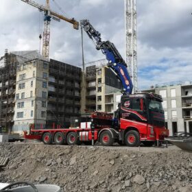 Punamusta kuorma-auto, jossa on sininen nosturi, työskentelee rakennustyömaalla. Taustalla näkyy keskeneräisiä kerrostaloja, rakennustelineitä ja useita nostokurkia. Maassa on kiviainesta, ja taivaalla pilvet vaihtelevat kirkkaasta valkoisesta tummempiin sävyihin. Ajoneuvo on pysäköity epätasaiselle sorapinnalle.